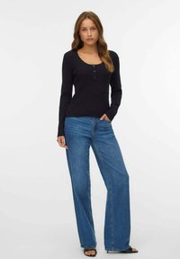 Donna in piedi con una mano in tasca, indossando un maglione nero a maniche lunghe con bottoni e jeans blu a gamba larga, di fronte a uno sfondo bianco.