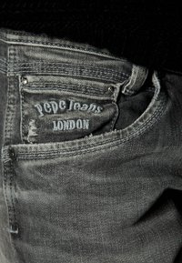 Mörkgrå denimjeans med en urtvättad textur, med en sydd etikett som säger "PGDC Jeans London" på bakfickan.