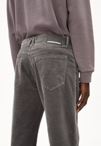 Persoon draagt grijze ribfluwelen broek met achterzakken en een zwart "ARMEDANGELS" label, gecombineerd met een zachtpaarse longsleeve.
