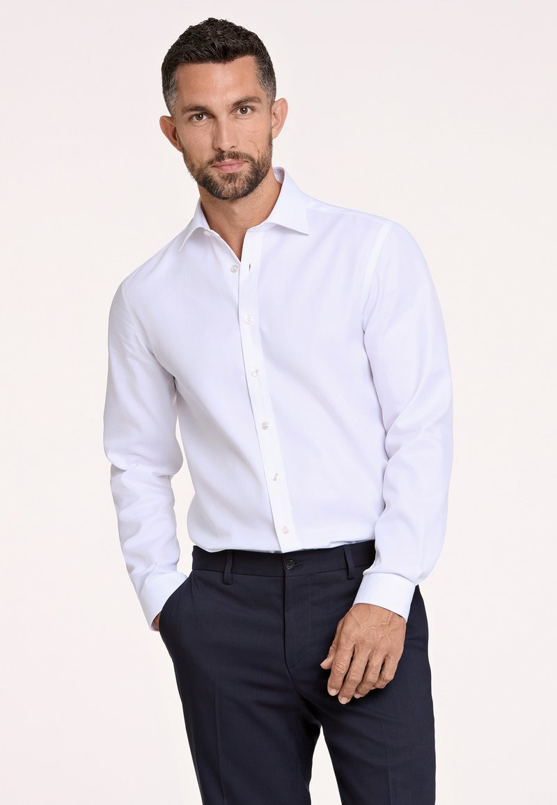 Camisa blanca de botón con cuello clásico, mangas largas y una textura suave; combinada con pantalones de vestir oscuros.