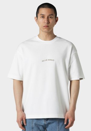 T-shirt en coton blanc à coupe décontractée, à manches courtes, col ras du cou, arborant un petit texte brodé "PAS DE MONACO" sur la poitrine.