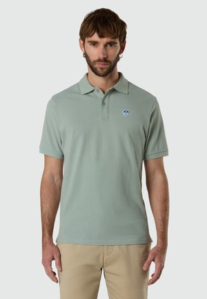 Uomo in piedi, rivolto verso l'obiettivo, indossa una polo a maniche corte verde chiaro con un piccolo logo blu e pantaloni beige su sfondo grigio chiaro.