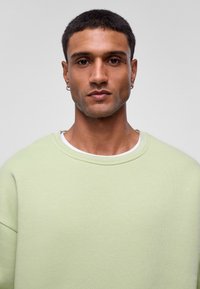 Lichtgroene sweatshirt met een ontspannen pasvorm en ronde halslijn. Heeft een zachte textuur en verlaagde schoudernaden. Gelaagd over een wit shirt.