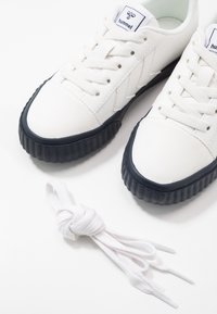 Baskets basses blanches avec semelles noires, lacets blancs attachés à côté, et un logo "hummel" sur la languette.