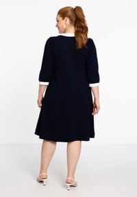 Navyblaues Kleid mit dreiviertel Ärmeln und weißen Manschetten, knielang, aus weichem Stoff und mit ausgestellter Silhouette.