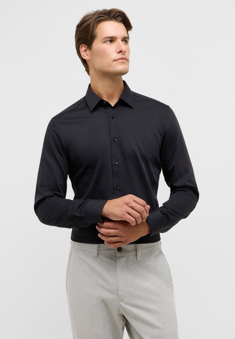 Camicia nera a maniche lunghe con bottoni e colletto classico, tessuto liscio e patta frontale. Abbinata a pantaloni grigi con texture.