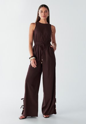 Jumpsuit sans manches marron avec une taille cintrée et des pantalons à jambes larges. Présente des liens à cordon aux chevilles et des accents dorés. Texture lisse.