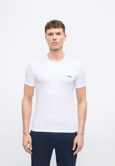 Νεαρός άνδρας με κοντά σγουρά μαλλιά, που φοράει εφαρμοστή λευκή μπλούζα με μικρό λογότυπο στο στήθος και σκούρο παντελόνι, μπροστά σε απλό φόντο.