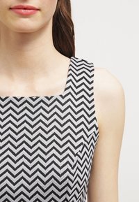 Svartvit tyg med chevronmönster på en topp med fyrkantig halsringning och breda band, som visar en slät textur och ren design.