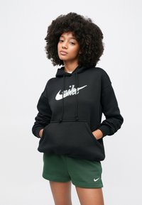 Μαύρο φούτερ Nike με κορδόνια και τσέπη μπροστά, με μεγάλο λευκό λογότυπο, συνοδευόμενο από πράσινο σορτς με μικρό λευκό λογότυπο.