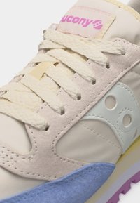 Κλείσιμο ενός παπουτσιού Saucony που δείχνει κορδόνια κρεμ, μπεζ σουέτ, γαλάζιες λεπτομέρειες και ροζ λογότυπο στη γλώσσα.