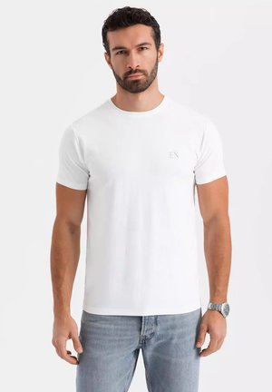 T-shirt bianco in cotone con collo rotondo e maniche corte. Presenta un logo grigio "EX" discreto sul lato sinistro del petto. Abbinato a jeans azzurri chiari.