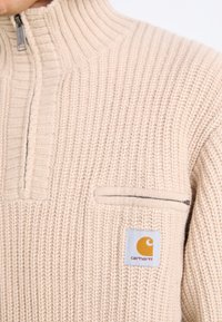 Bežni pleteni zip-up pulover z visokim ovratnikom, ki ima majhen žep na prsih, rebrasto teksturo in oranžno oznako Carhartt.
