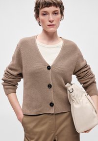 Cardigã castanho com nervuras e decote em V, adornado com três botões pretos, usado sobre uma blusa creme. Acompanhado por uma mala creme e calças caqui.