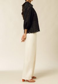 Blusa nera ricamata con maniche a sbuffo e orlo scallop, abbinata a pantaloni bianchi a gamba larga e sandali arancioni con tacco.