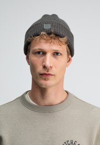 Grijze gebreide beanie met een geribbelde textuur en een klein vierkant logo-opdruk. Draagt bij een lichtgrijze sweatshirt met tekst en een ronde halslijn.