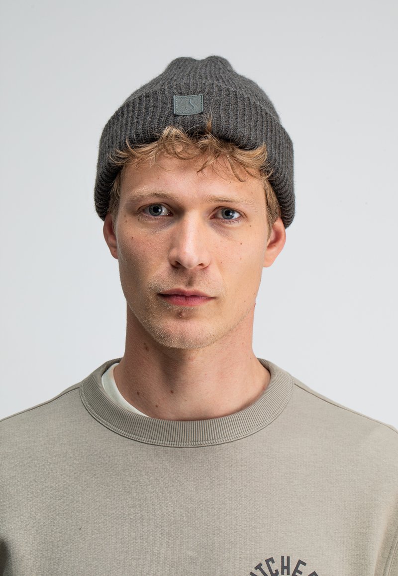Grijze gebreide beanie met een geribbelde textuur en een klein vierkant logo-opdruk. Draagt bij een lichtgrijze sweatshirt met tekst en een ronde halslijn.