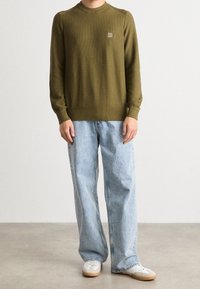 Pull en maille vert olive à col rond et accents d'épaule, associé à un jean large bleu clair et des baskets blanches.