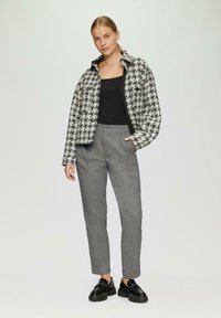 Schwarze und weiße Hahnentrittmuster-Jacke, taillierter Schnitt, über einem schwarzen Oberteil; kombiniert mit grauen, maßgeschneiderten Hosen und schwarzen Slippers, glatte Textur.