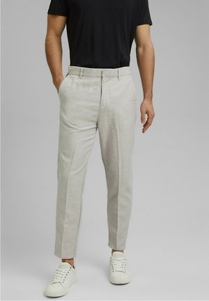 Mężczyzna ubrany w jasnobeżowe spodnie na miarę, białe sneakersy i czarną koszulę z krótkim rękawem, stojący z jedną ręką w kieszeni.