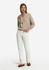 Beige Langarmshirt mit einer Schleifendetail am Hals, kombiniert mit weißen gerade geschnittenen Jeans und braunen Ballerinas, präsentiert ein lässiges Outfit.