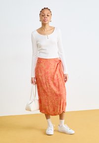 Hvit ribbet langermet topp, rød-oransje innpakningsskjørt med blomsterdesign, hvite joggesko og en liten hvit håndveske, posert mot en nøytral bakgrunn.