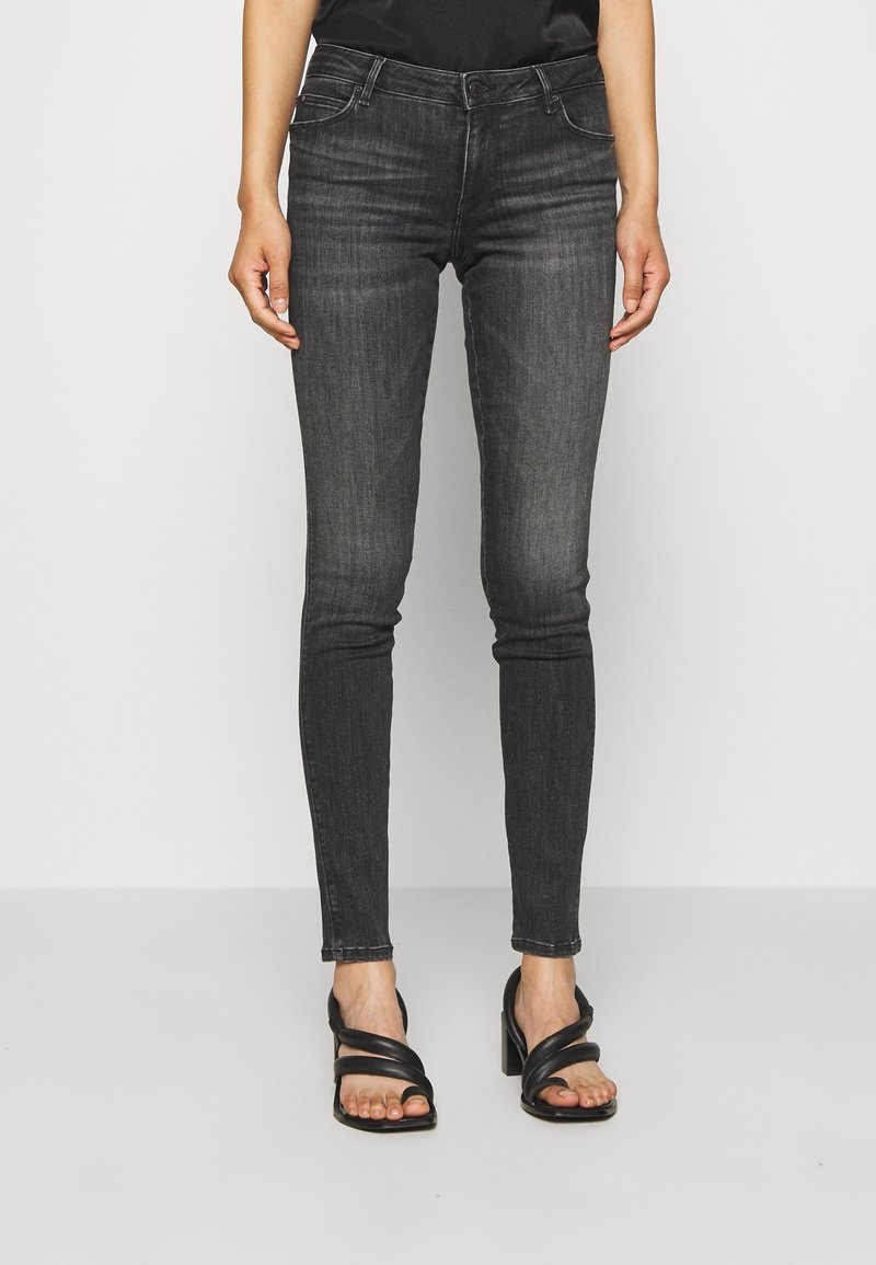 Jeans negros ajustados con un acabado desgastado, que presentan un diseño clásico de cinco bolsillos y costuras en contraste. Llevados con sandalias de tacón negras.