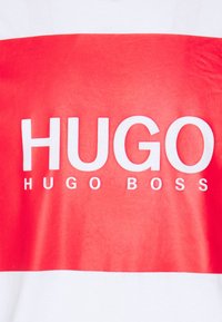 Bílý tričko s velkým červeným obdélníkovým grafickým prvkem, na kterém je nahoře tučný bílý text "HUGO" a pod ním menší "HUGO BOSS". Hladká bavlněná tkanina.