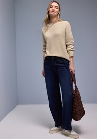 Femme portant un pull en maille beige, un jean large bleu foncé et des baskets blanches, tenant un sac fourre-tout matelassé marron sur un fond gris uni.