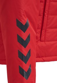 Rode pufferjas met een gewatteerde textuur, voorzien van grote zwarte chevronaccenten op de linker manchette voor een design-detail.