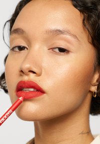 Red lip liner is applied to the lips. Skin is smooth with a natural glow. Hair is curly, framing the face. Simple background.