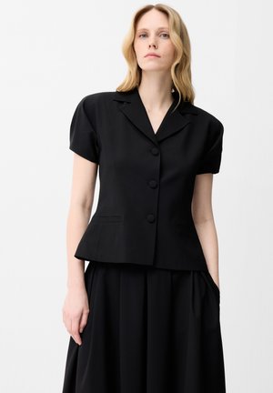 Kvinde med lyst blond hår iført kortærmet sort knapblazer og matchende sort plisseret nederdel mod en ensfarvet lys baggrund.