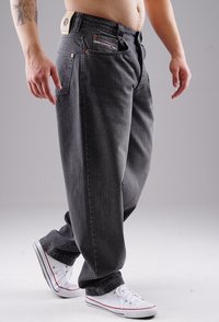 Dunkelgraue Denim-Hosen mit lockerer Passform, fünf Taschen und kontrastierenden Nähten. Getragen mit weißen Sneakern. Subtile Textur sichtbar.
