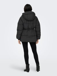 Black puffer jacket with a hood, quilted design, elastic waistband, cinched back detail, and worn over black leggings and ankle boots.