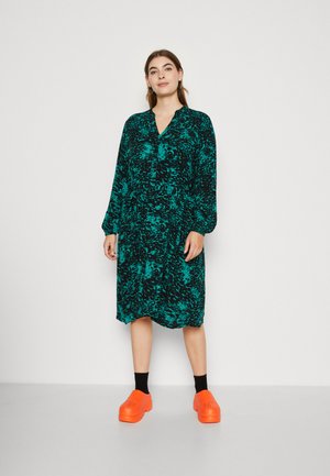 Abito verde e nero a fantasia con maniche lunghe e scollatura a V, abbinato a scarpe con tacco arancioni vivaci e calze nere.