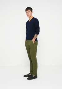 Pull bleu marine à col en V, pantalon tapered vert olive et chaussures noires à enfiler. Le mannequin se tient avec les mains dans les poches devant un fond uni.