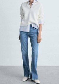 Suéter blanco acanalado sobre una camisa de cuello azul claro, combinado con jeans de campana azul claro y zapatos slip-on blancos.
