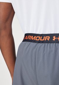 Graue Sportshorts aus strukturiertem Stoff mit einem schwarzen Bund, der das "ARMOUR"-Logo in Orange zeigt.