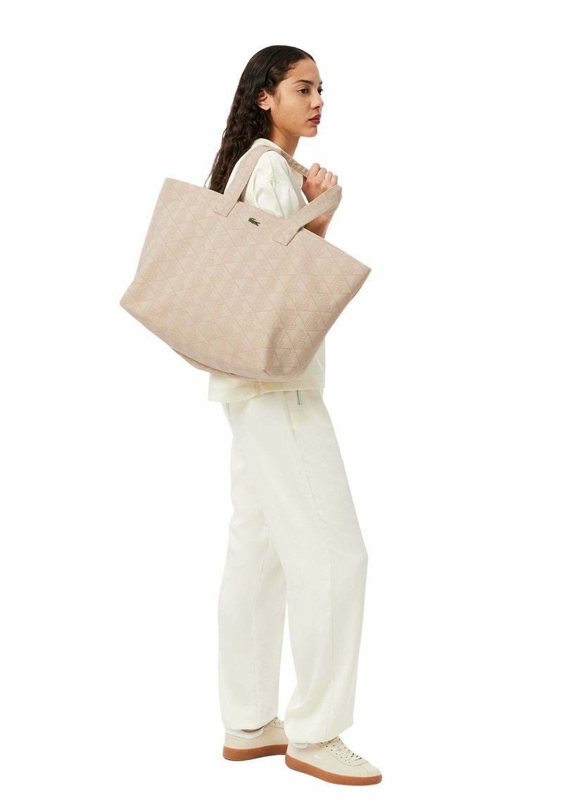Frau in weißem Outfit trägt große beige Tragetasche mit strukturiertem Dreiecksmuster und Logo auf der Schulter, steht vor weißem Hintergrund.