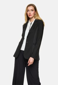 Schwarze taillierte Jacke mit Revers, zwei Knöpfen und seitlichen Taschen. Sie hat einen strukturierten Stoff und eine lockere Passform. Über einem weißen Oberteil getragen.