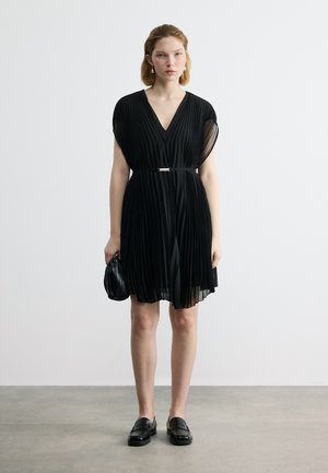 Femeie purtând o rochie neagră plisată până la genunchi, cu o curea, ținând o poșetă neagră mică, stând pe un podea gri, în fața unui perete alb simplu.