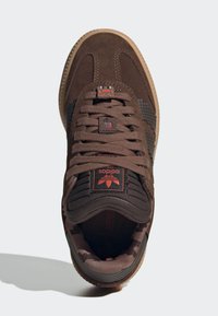 Brauner Sneaker aus Wildleder und Mesh-Materialien, mit pixeliertem Design, roten Akzenten, strukturierten Schnürsenkeln und einer Gummi-Außensohle.