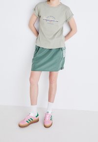 Ljust grön t-shirt med upprullade ärmar och ett grafiskt tryck, kombinerad med en grön kjol med sidofickor och vita ränder, samt rosa skor.