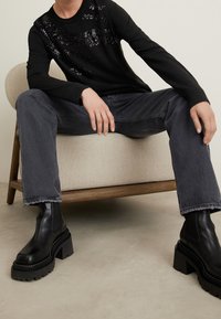 Schwarzer Pullover mit Paillettenakzenten, graue Jeans mit ausgefransten Säumen und klobige schwarze Stiefeletten, sitzend auf einem hell bezogenen Stuhl.