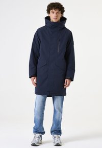 Marineblaue isolierte Parka mit hohem Kragen, Reißverschluss und Druckknopfverschluss, zwei vorderen Taschen und verstellbarem Kapuze. Lässiger Schnitt; über Denim getragen.