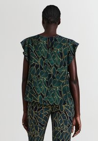 Donna con i capelli intrecciati che indossa un top e pantaloni neri abbinati con motivi di foglie tropicali verdi e gialle, vista da dietro.