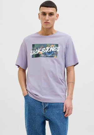 Lavendel T-shirt med korte ærmer, prydet med et grafisk tryk af tropiske planter og fed tekst. Bæret sammen med afslappede blå denim.