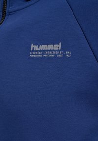 Modrá športová mikina s hladkou textúrou. Obsahuje viditeľný svetlo sivý potlač loga s nápisom "hummel" a textom v modernom písmene.