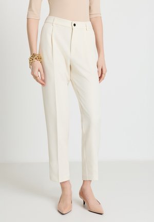 Femme portant un pantalon crème taille haute sur mesure, des talons pointus couleur chair, un haut beige, avec des bracelets et des bagues en or aux mains sur les côtés.