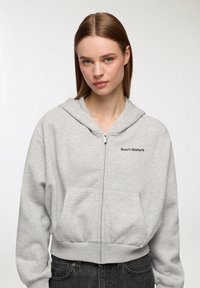 Szary rozpinany hoodie z przednią kieszenią i napisem "Nie przeszkadzać". Posiada kaptur ze sznurkiem i ściągacz w pasie. Miękka tekstura materiału.